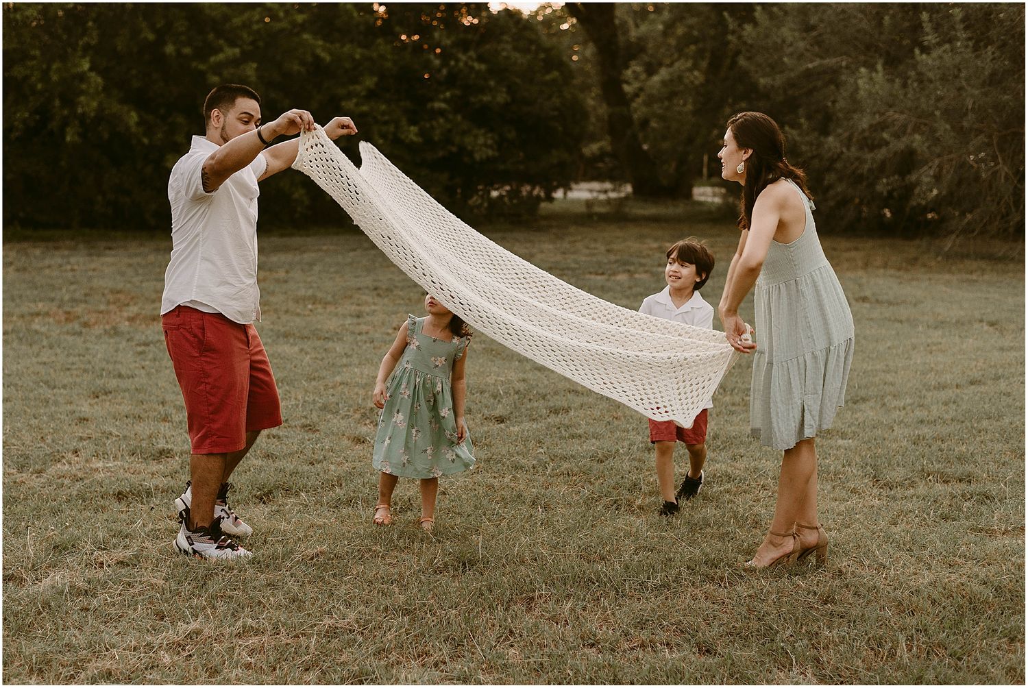A family plays together with a white sheet held up in the air on a grassy field.