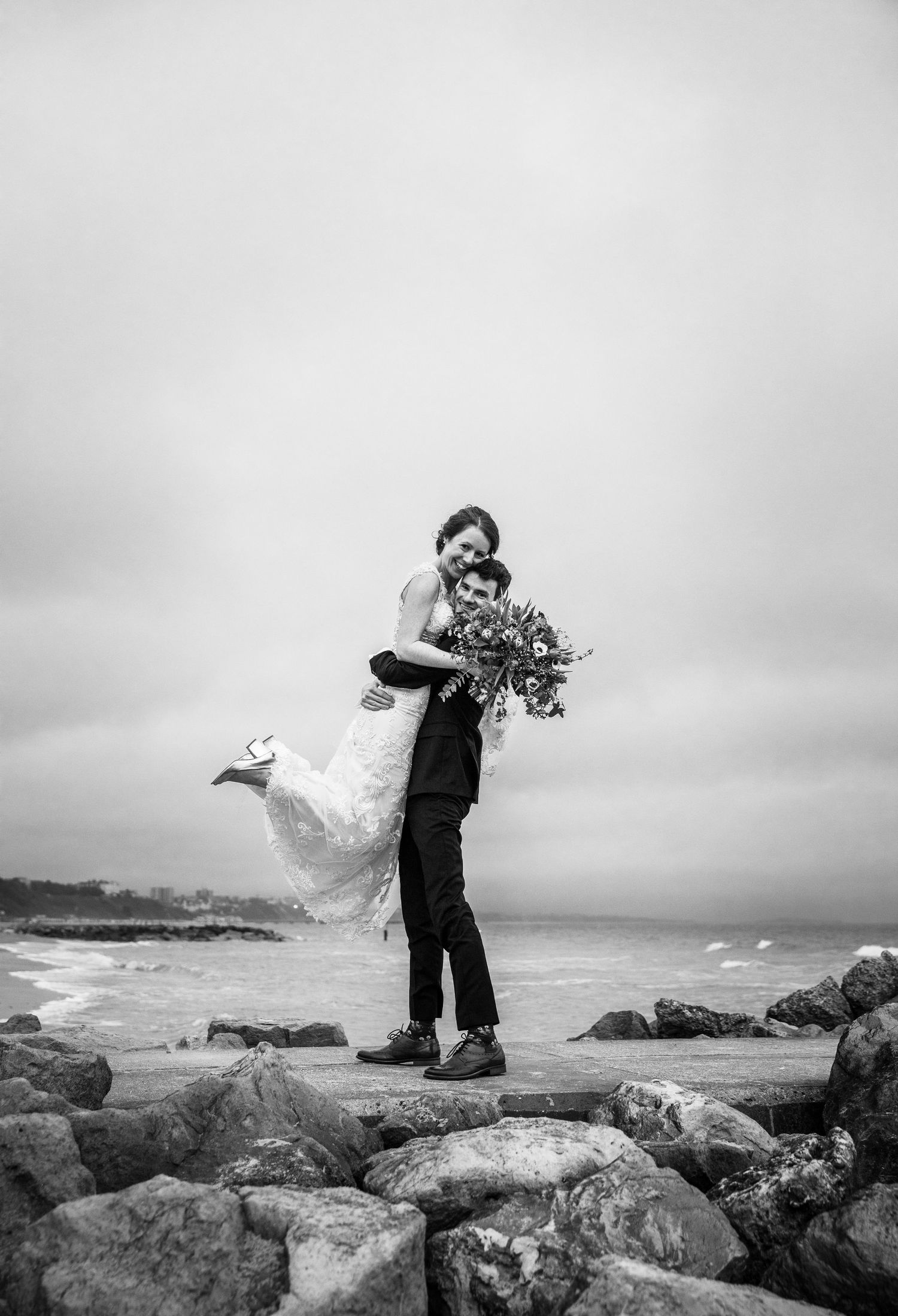 Romantic black and white portrait of couple embracing on rocky coastline.