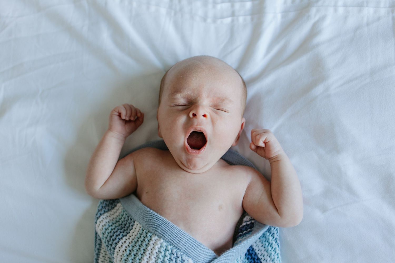 Newborn baby yawning while lying on white bedding wearing a blue knit blanket.