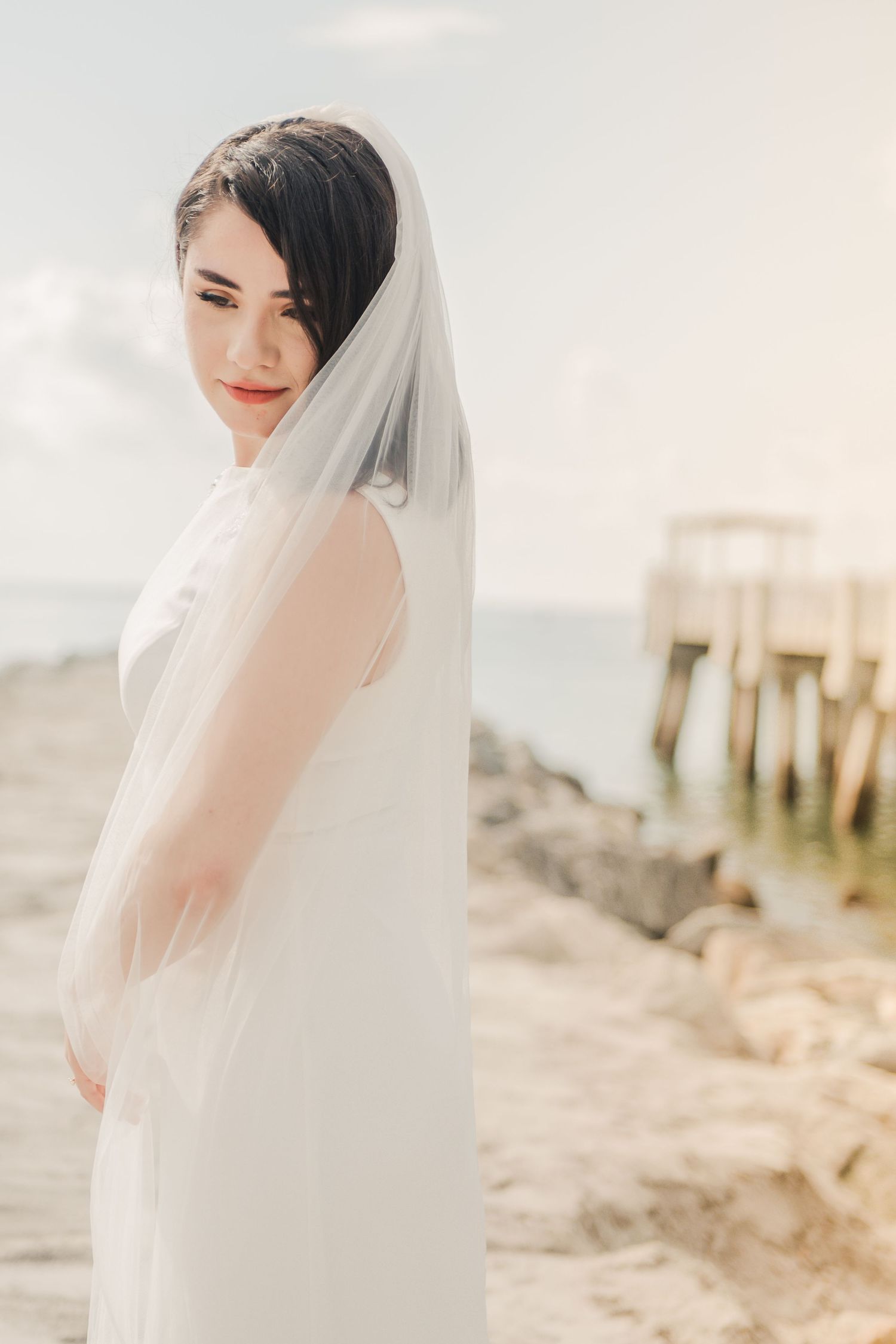 Elegant bride in a flowing white dress and veil poses serenely on a beach with a wooden pier in the background.