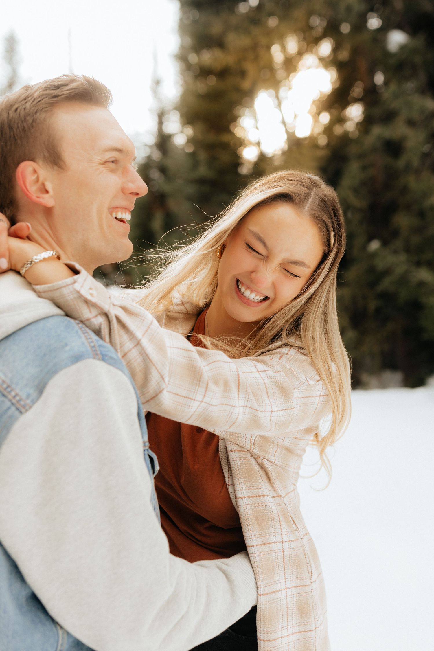 Woman hugging fiancé and laughing with the sun behind her in the trees