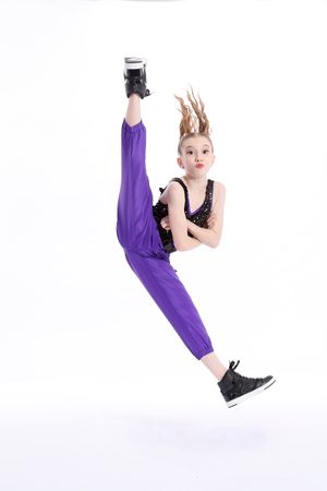 Dancer in purple pants and black top performs dramatic high kick in mid-air jump against white background.