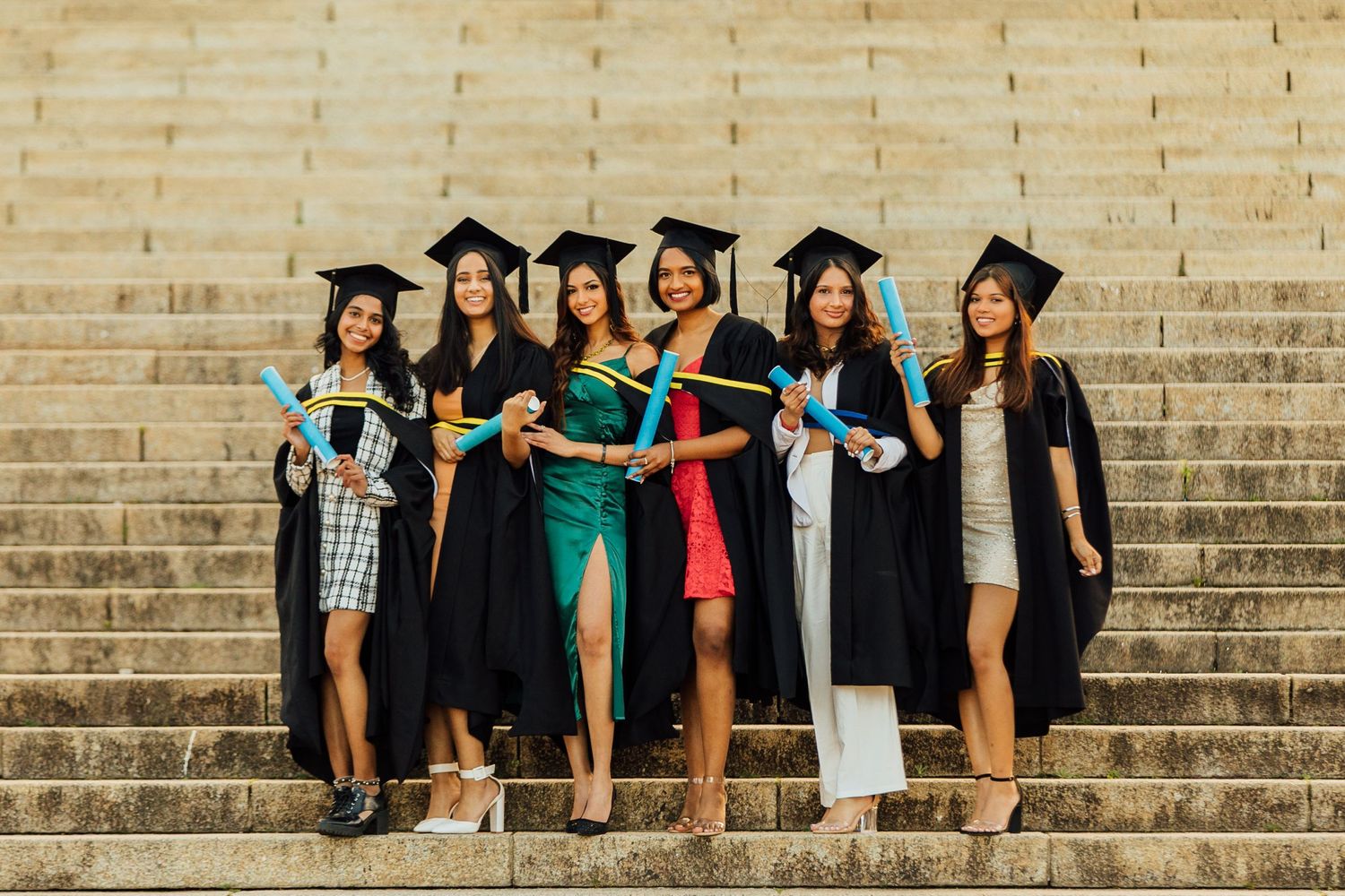 A group of graduates in caps and gowns stand together on stone steps holding diplomas during a sunny graduation ceremony.