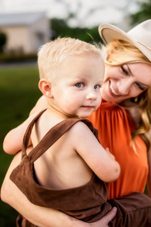 A toddler in suspenders in his Mom's arms looking at the camera.