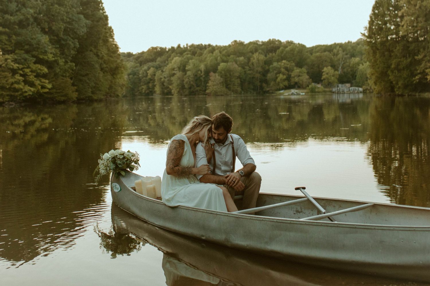 Couple sharing a romantic moment in a wooden rowboat on a serene lake surrounded by lush green trees.