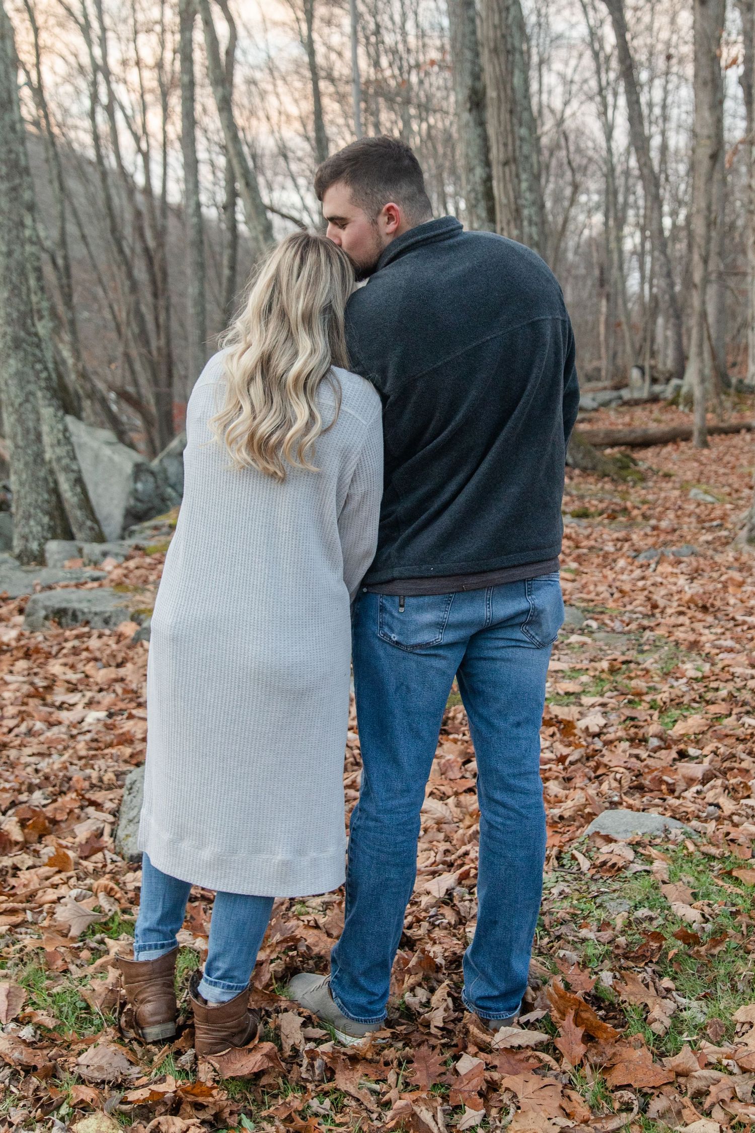 A couple in casual wear stands together on fallen autumn leaves in a wooded setting.