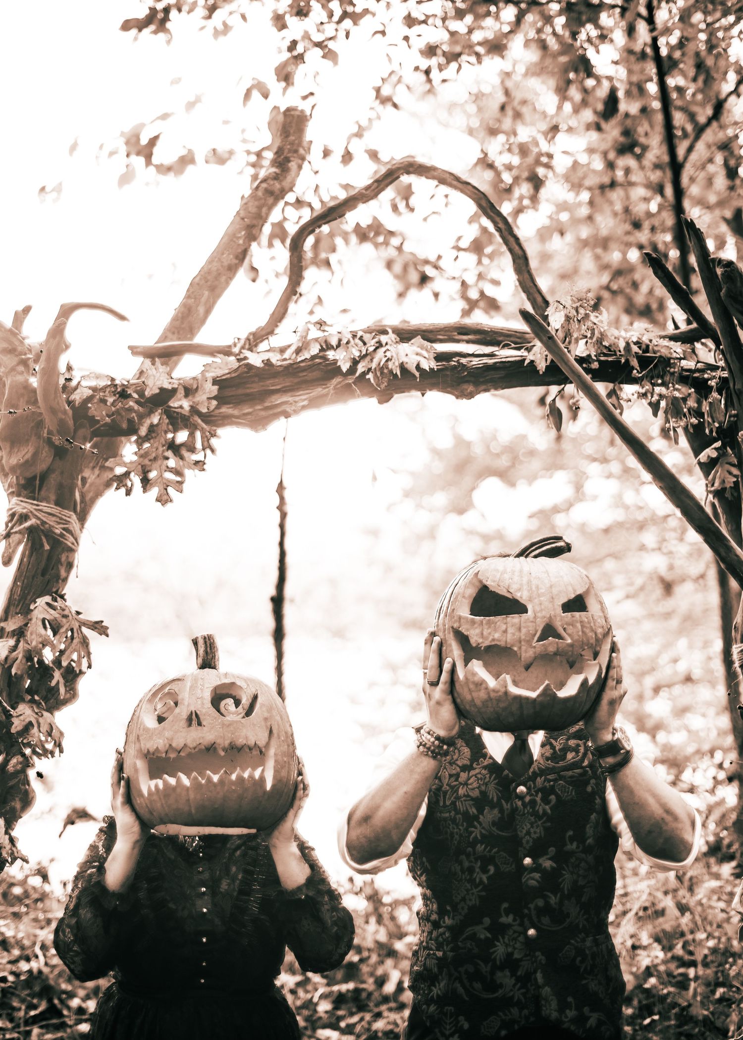 Two people wearing spooky jack-o-lantern masks pose under tree branches in a sepia-toned photo.