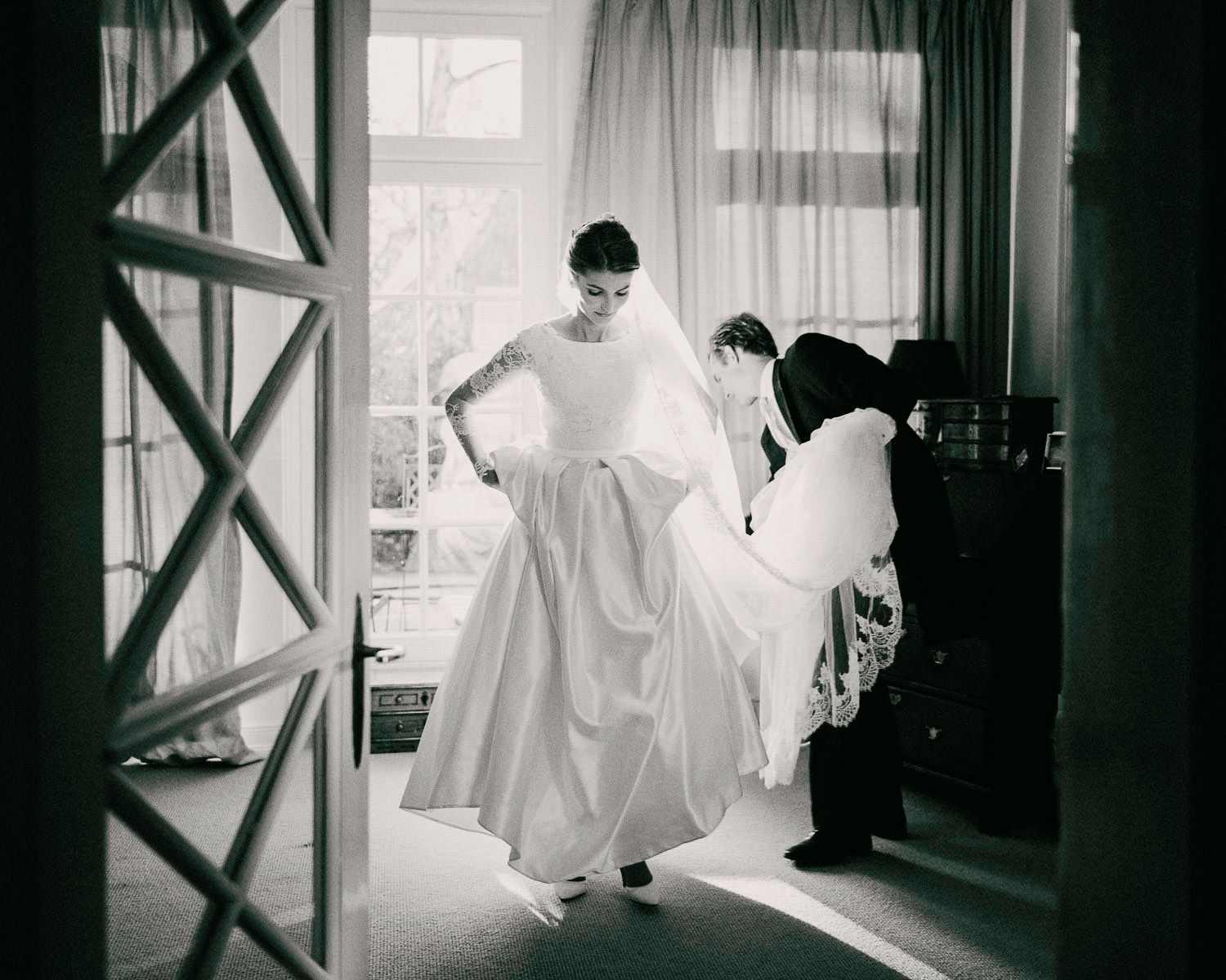 A black and white photo showing preparations before a wedding as attendants help with a dress.