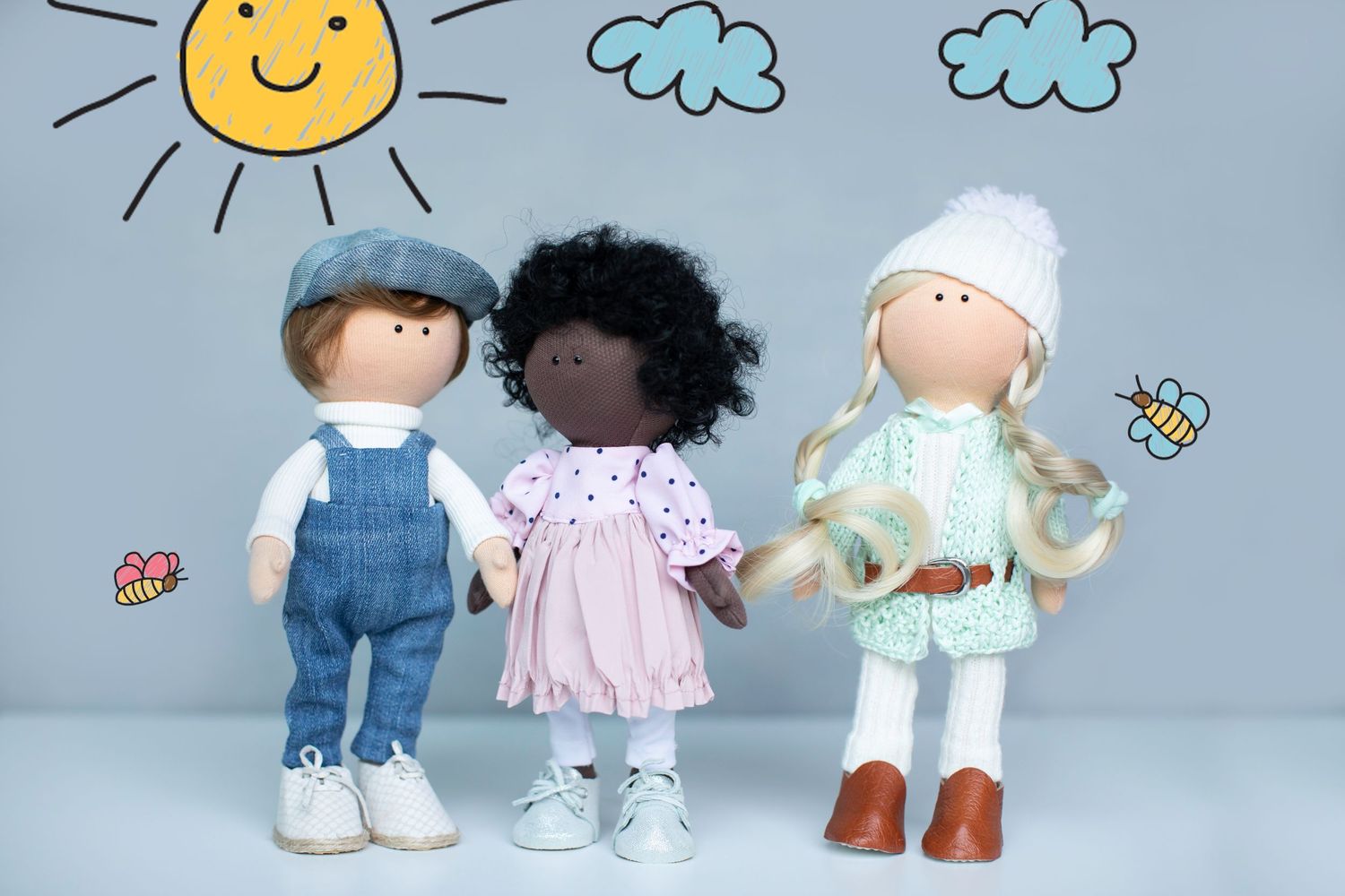 Three diverse dolls standing together against a light blue background with hand-drawn sun and clouds.