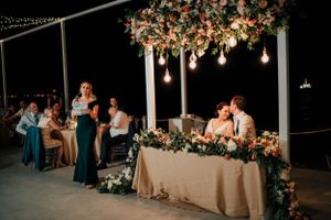 Wedding party at head table under floral arrangements and hanging Edison bulbs during reception.