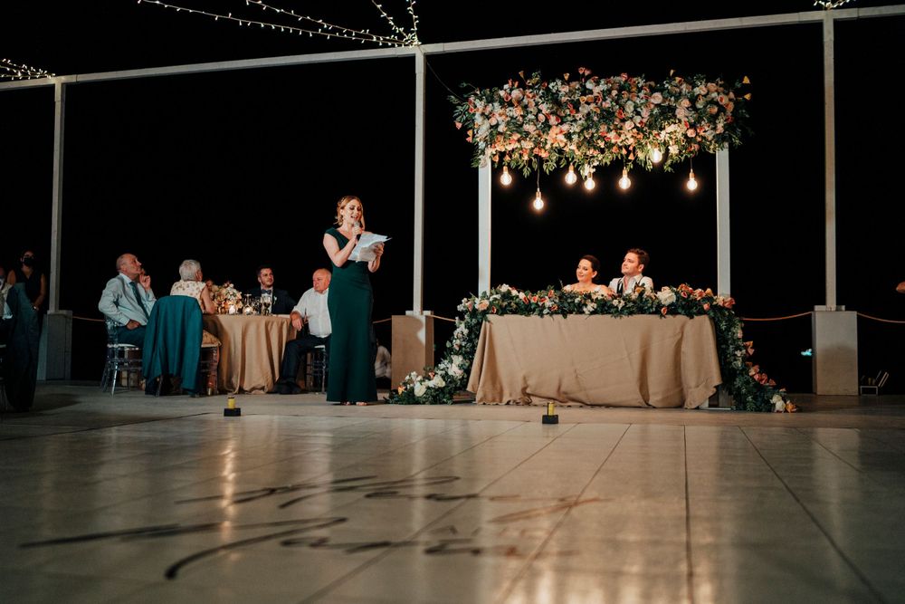Wedding reception setup with elegant floral chandelier and string lights during evening speeches.