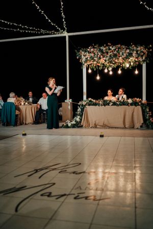 Evening wedding reception speech with decorative floral installation and projected text on dance floor.
