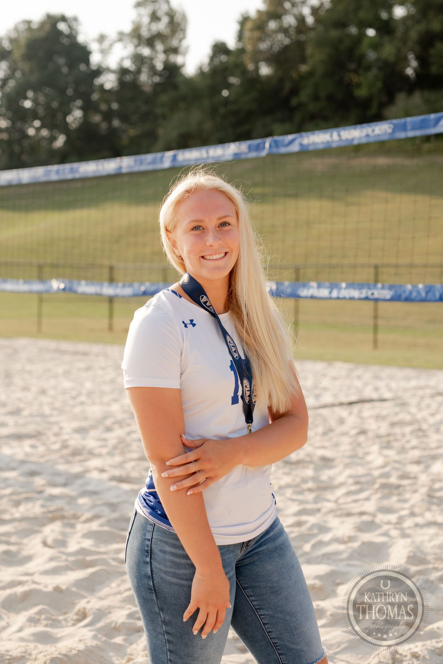 Athlete in white uniform smiles on sandy volleyball court during golden hour.
