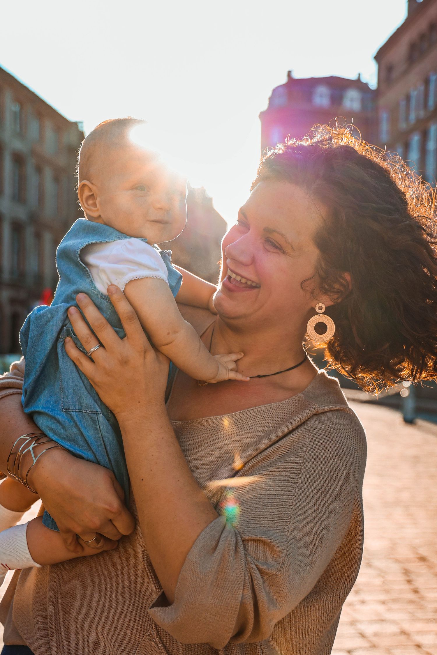 A baby in blue denim outfit is lifted joyfully against a sunlit urban backdrop.