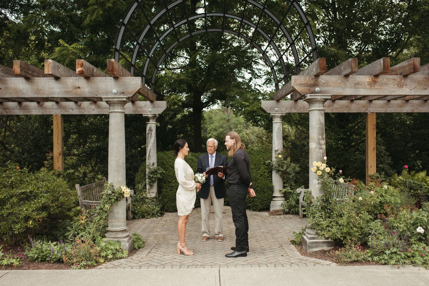 Intimate ceremony takes place under wooden pergola in lush garden setting.