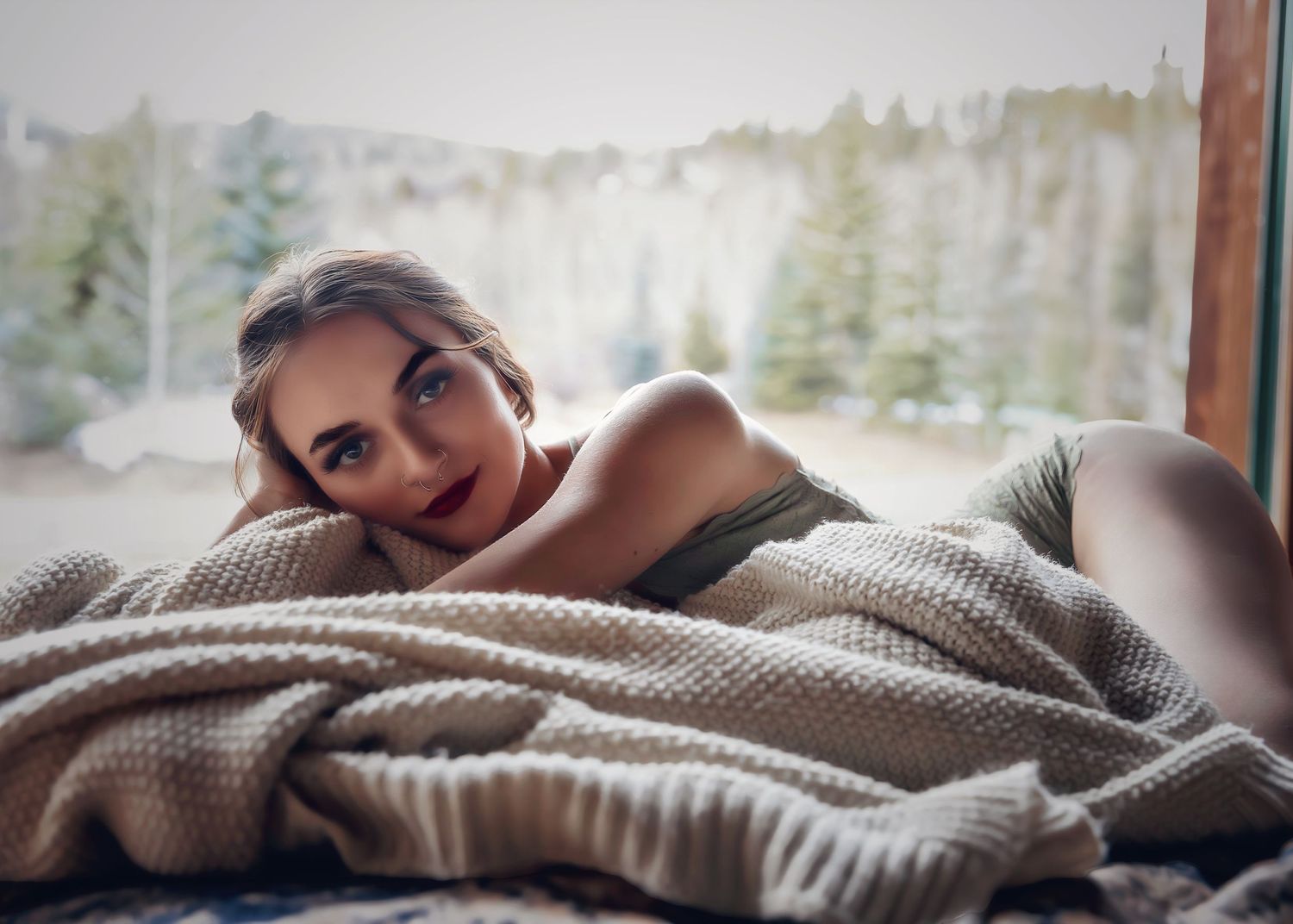 Blonde woman lying on bed wrapped in cozy knit blanket with snowy forest visible through window.