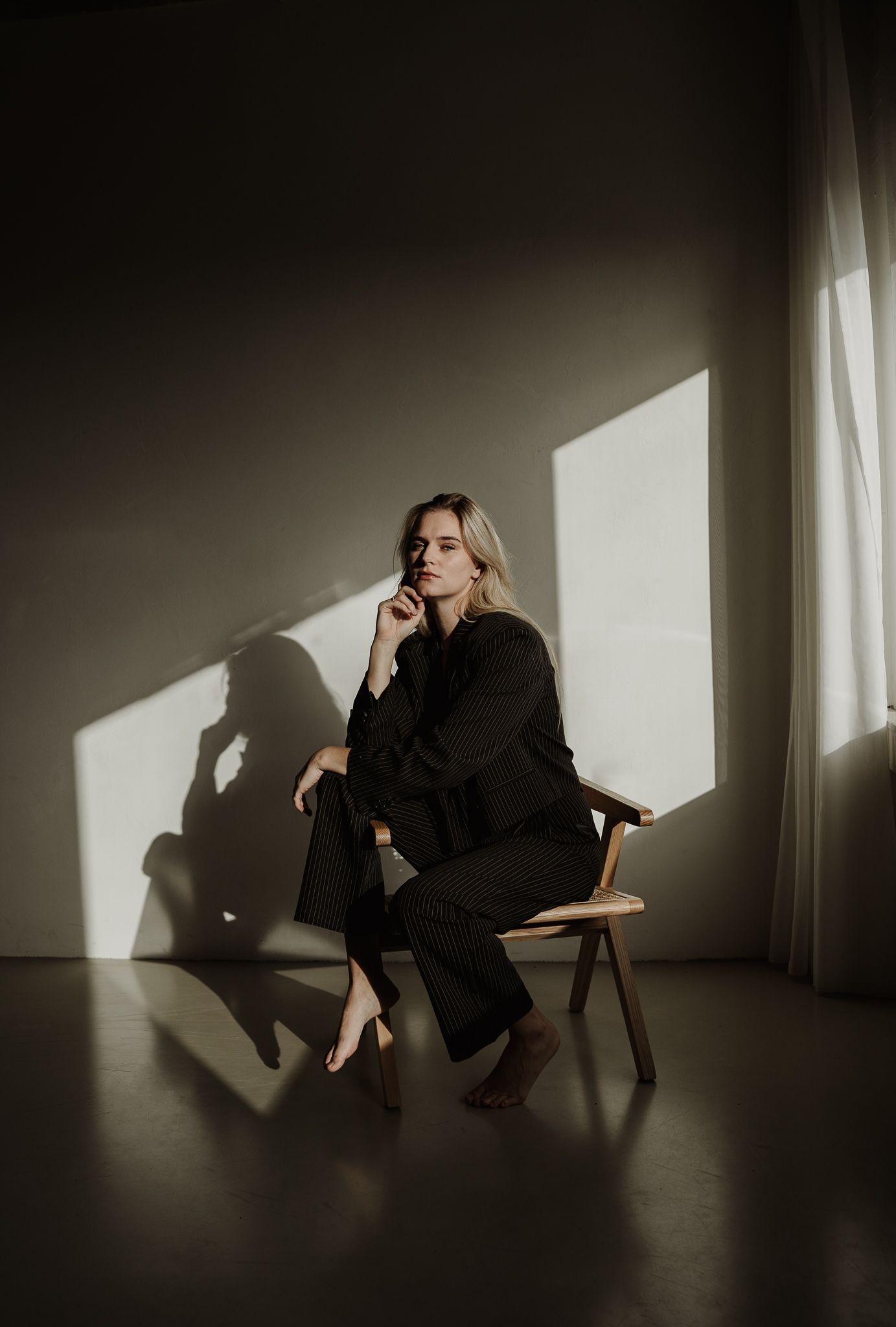 Moody portrait with dramatic shadows of person seated in wooden chair by window.