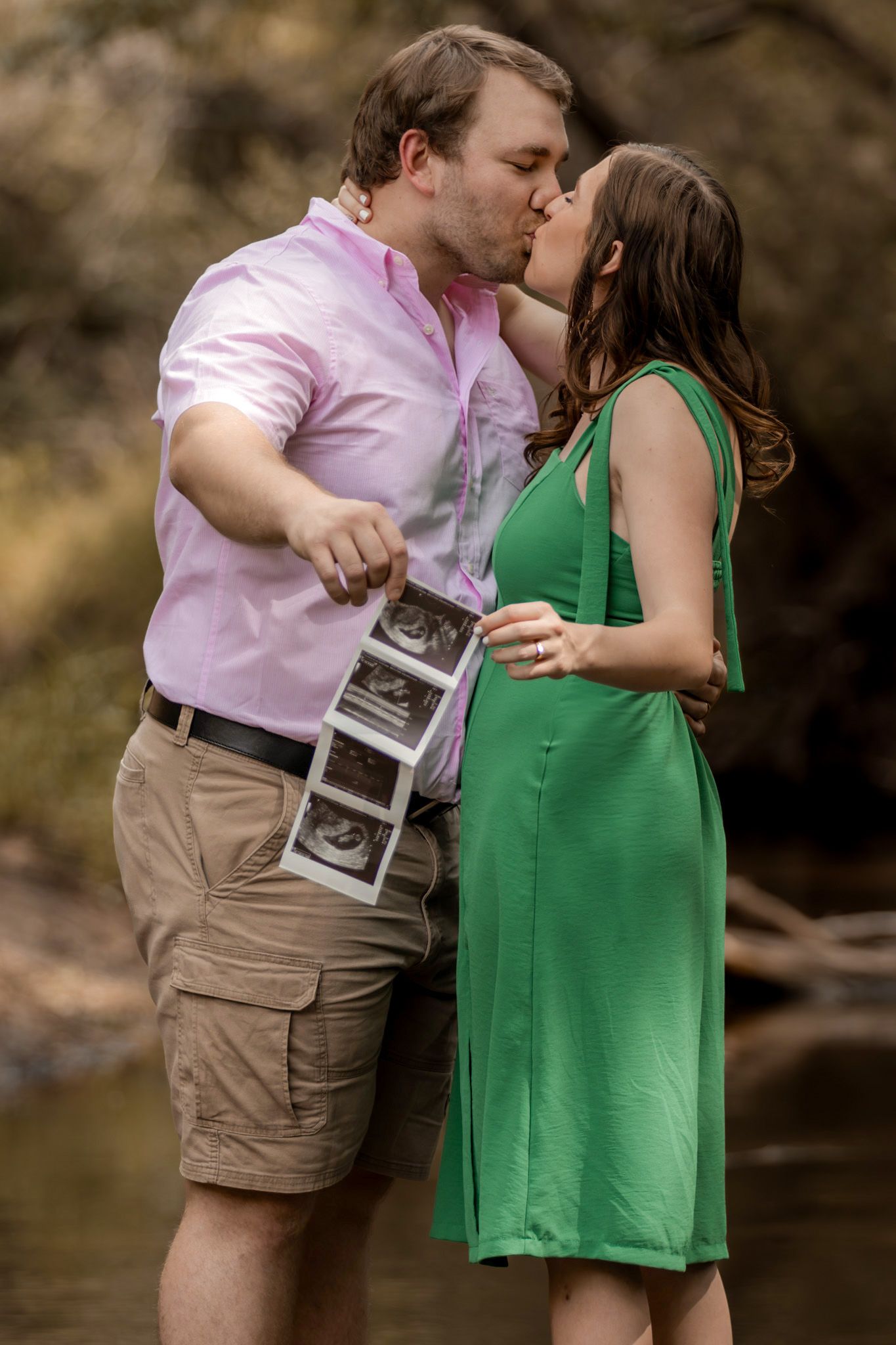 Couple shares a kiss while holding sonogram photos during pregnancy announcement photoshoot.