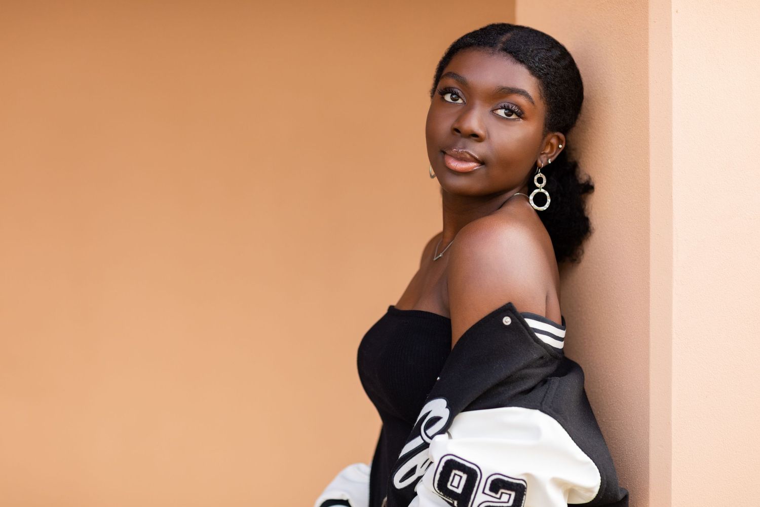 A high school senior against peach wall wearing black top and varsity jacket.