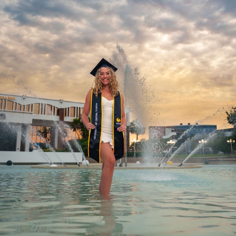 Graduate in cap and gown standing in fountain water during golden sunset.