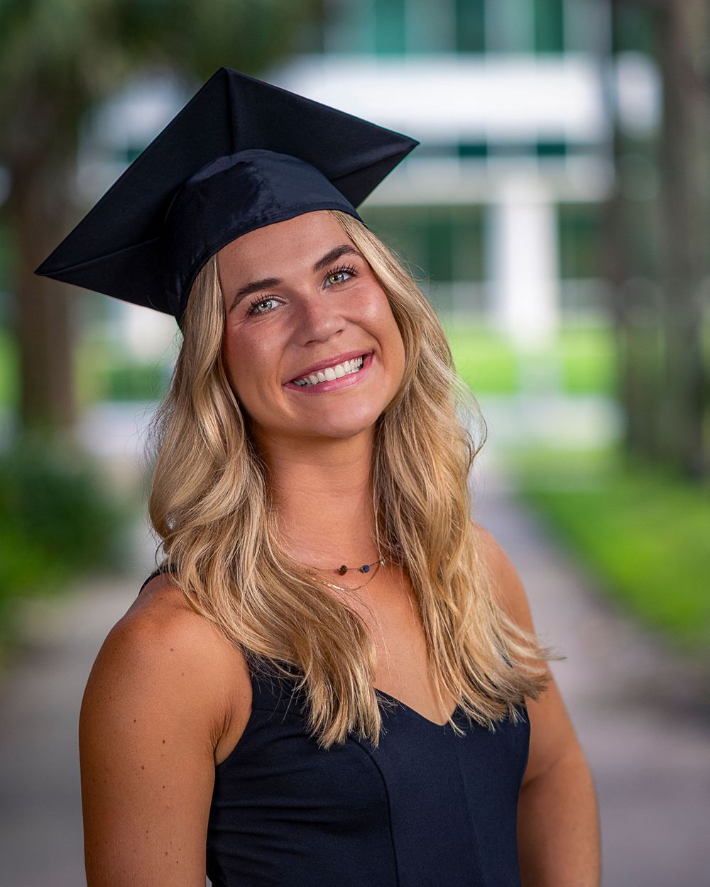 A graduate in cap and gown poses for a portrait on a tree-lined campus path.