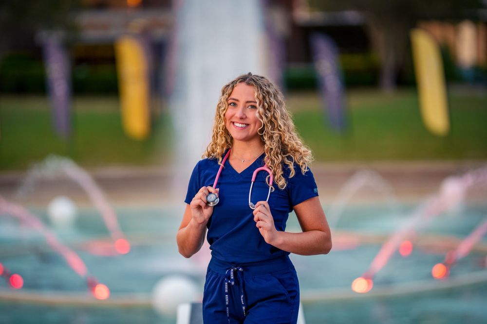Medical professional in blue scrubs outdoors near a fountain at sunset.