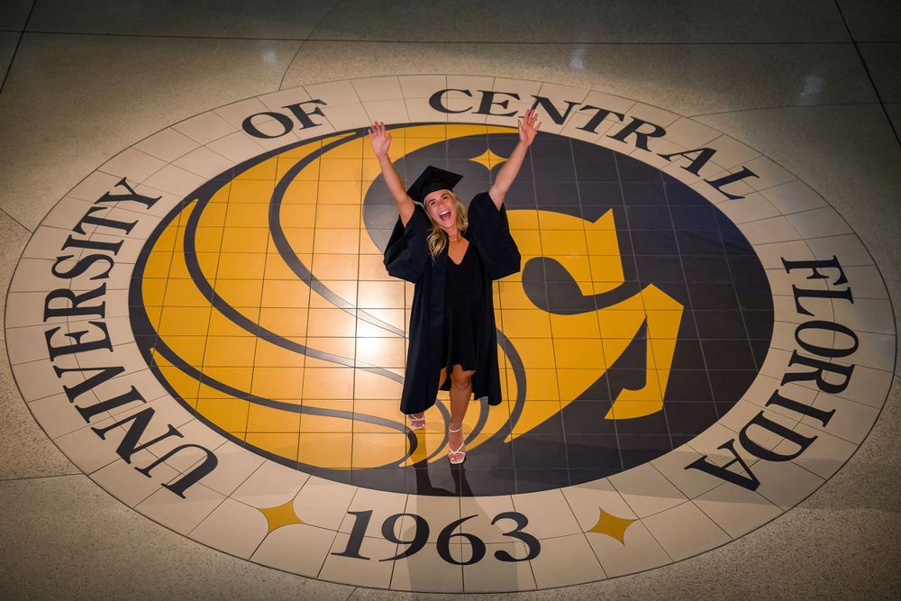 Graduate poses on University of Central Florida logo medallion embedded in floor.