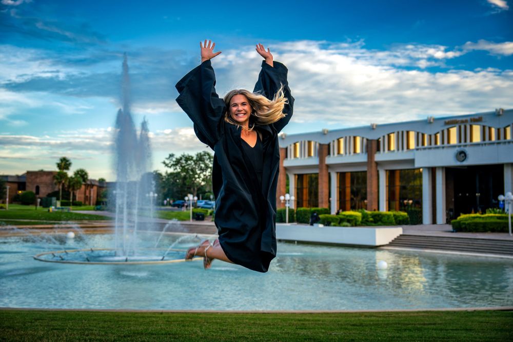 Graduate in black cap and gown jumping joyfully in front of a fountain and campus building.