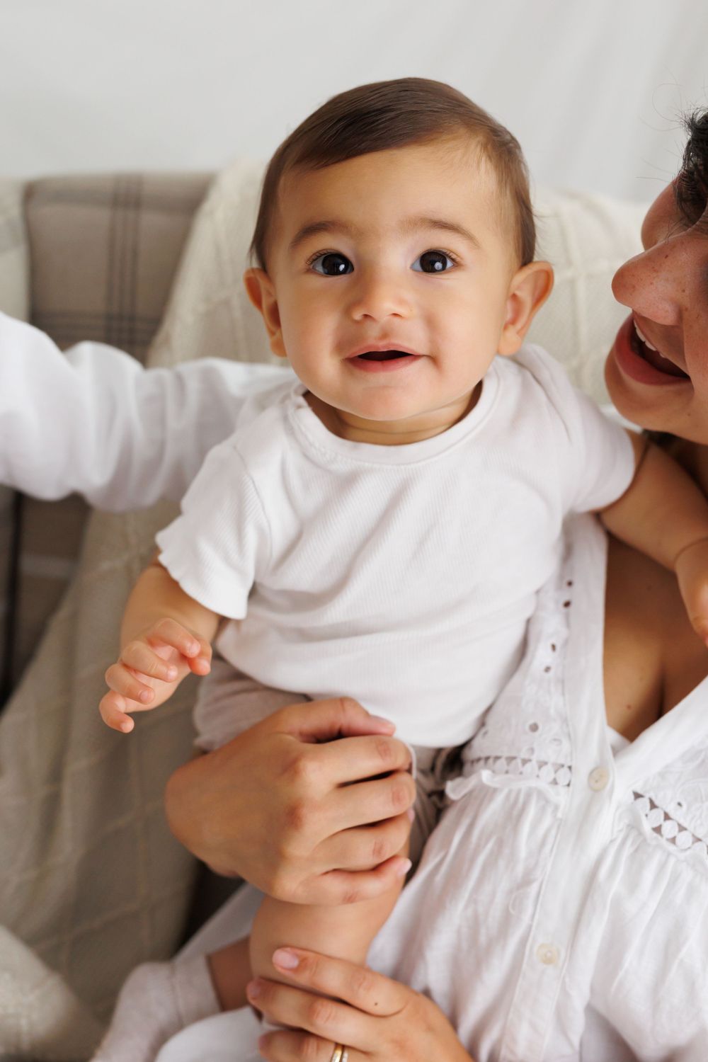 Happy baby in white outfit smiles during candid family photo moment.