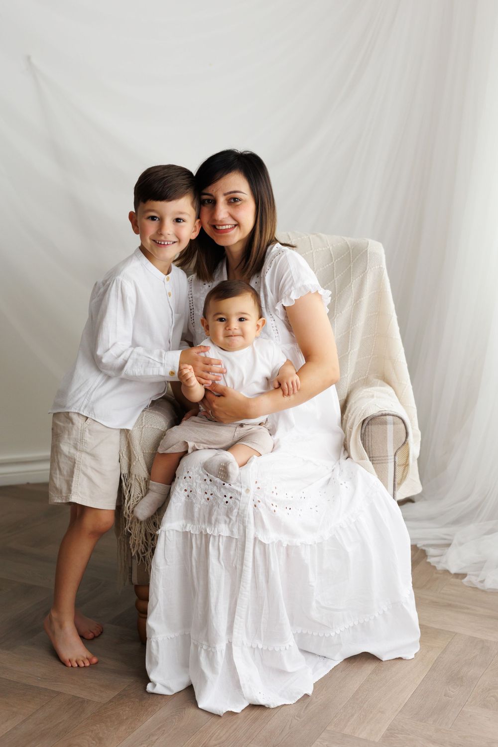 Family wearing coordinated white outfits poses together in armchair for portrait.
