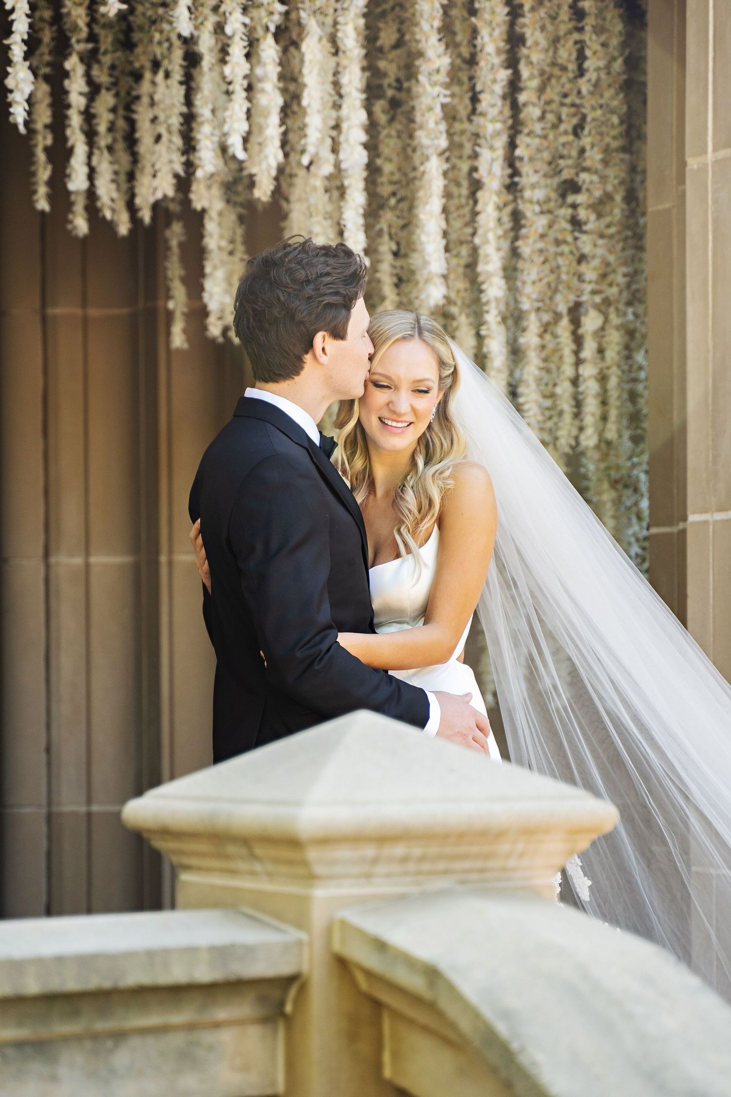 Romantic wedding couple embraces under hanging floral decorations.