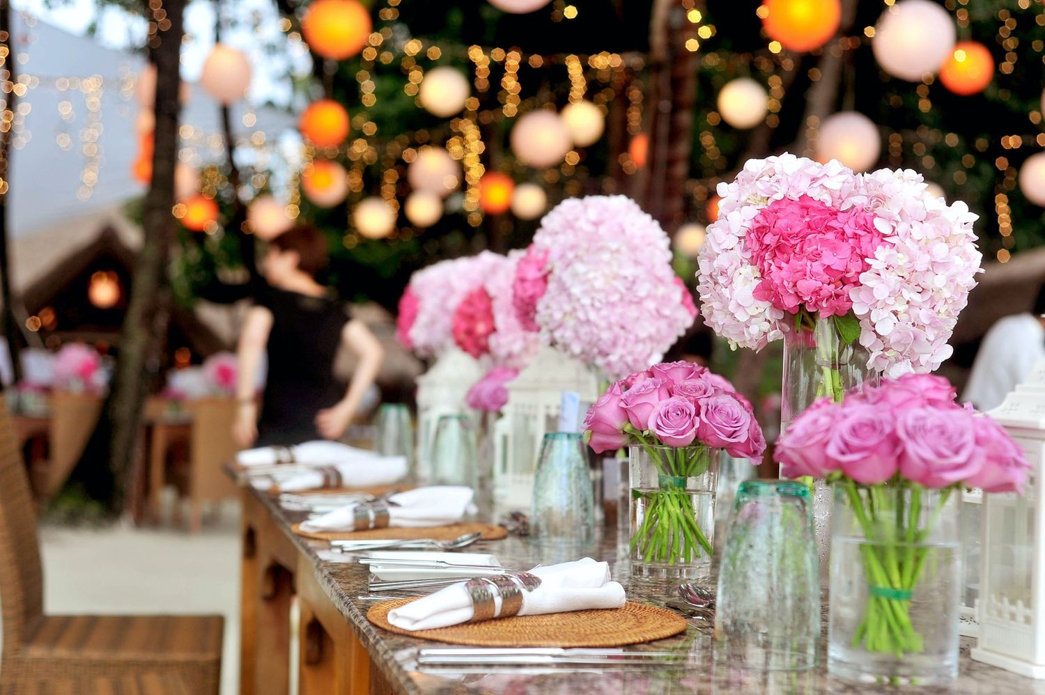 Elegant outdoor wedding reception table decorated with pink peony centerpieces and glowing lanterns overhead.
