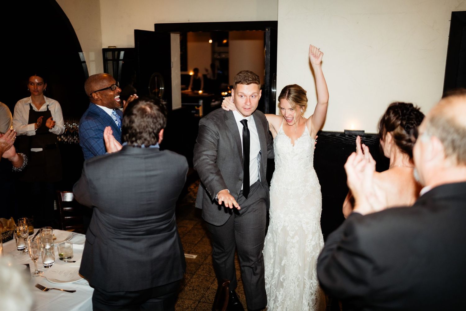 A wedding couple celebrates on the dance floor with their guests at an indoor reception venue.