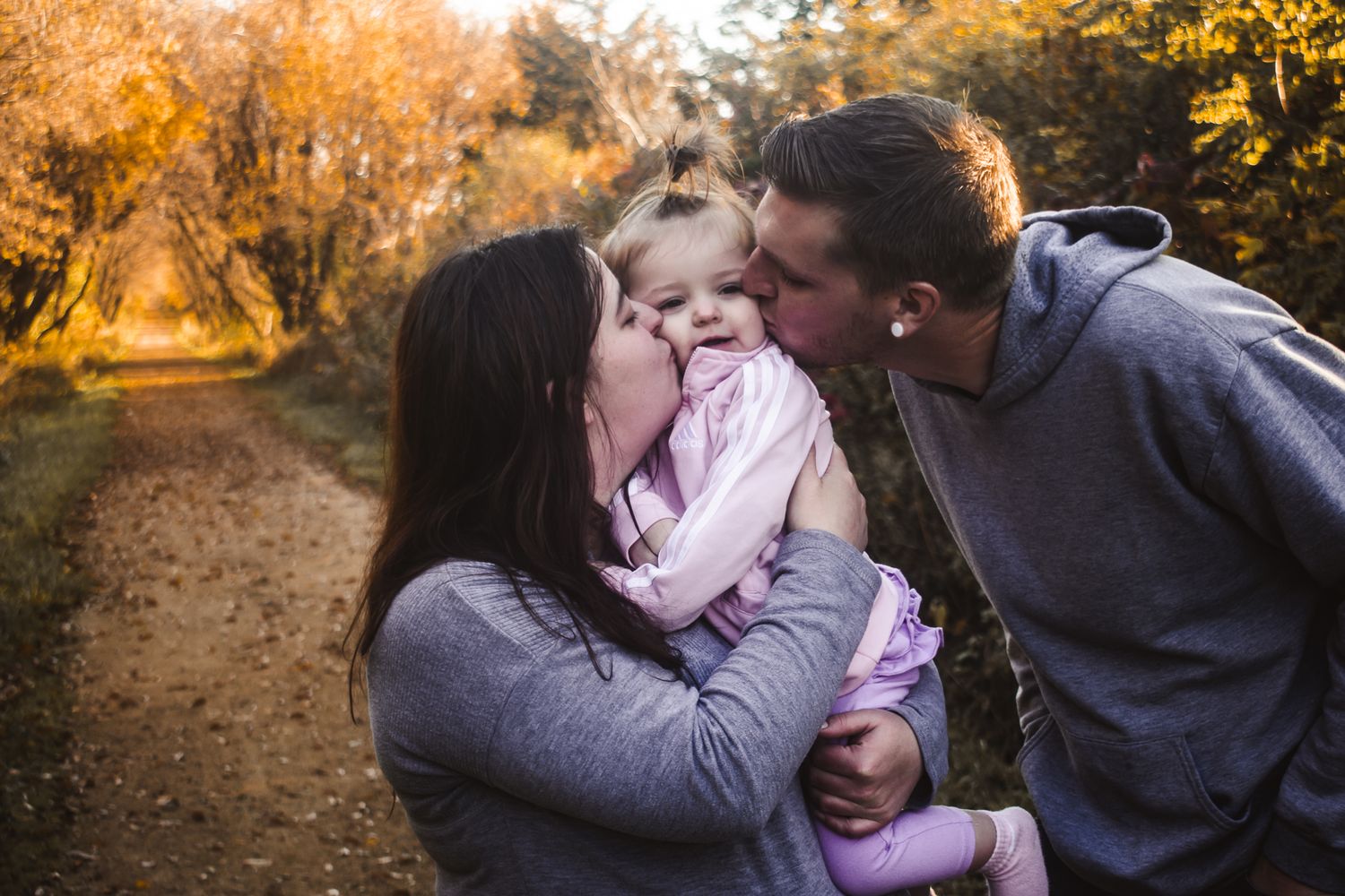 Parents kiss their young child on both cheeks during autumn outdoor photoshoot.