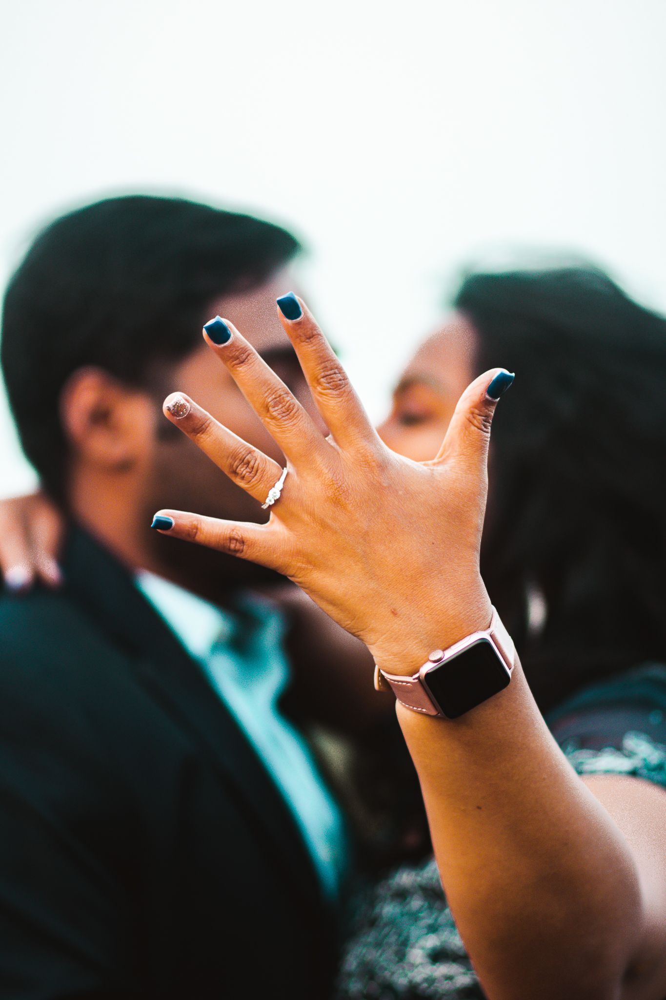 Close up of hand showing engagement ring with couple blurred in background.