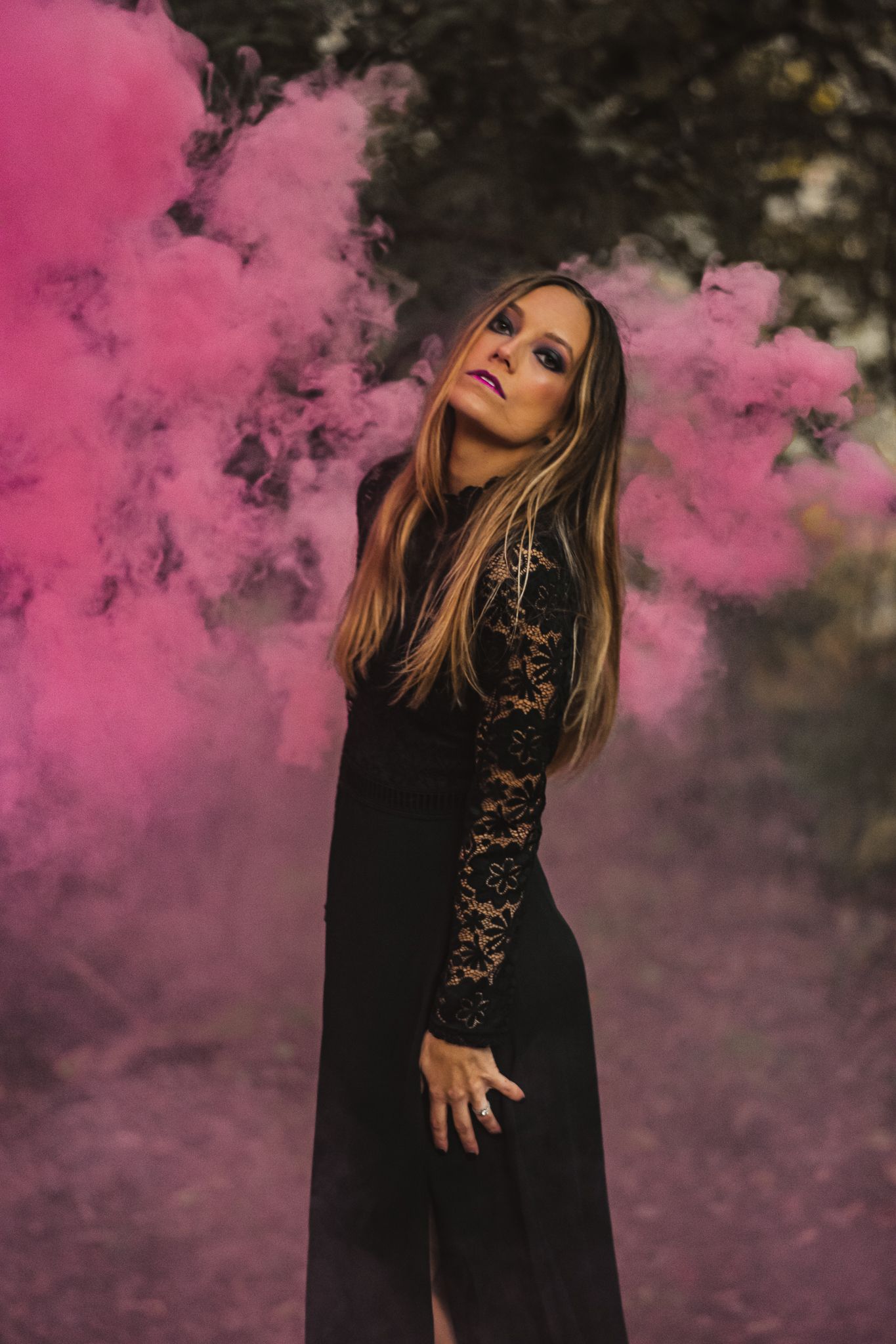A dramatic portrait with pink smoke effects showing a woman in black lace dress posing outdoors.