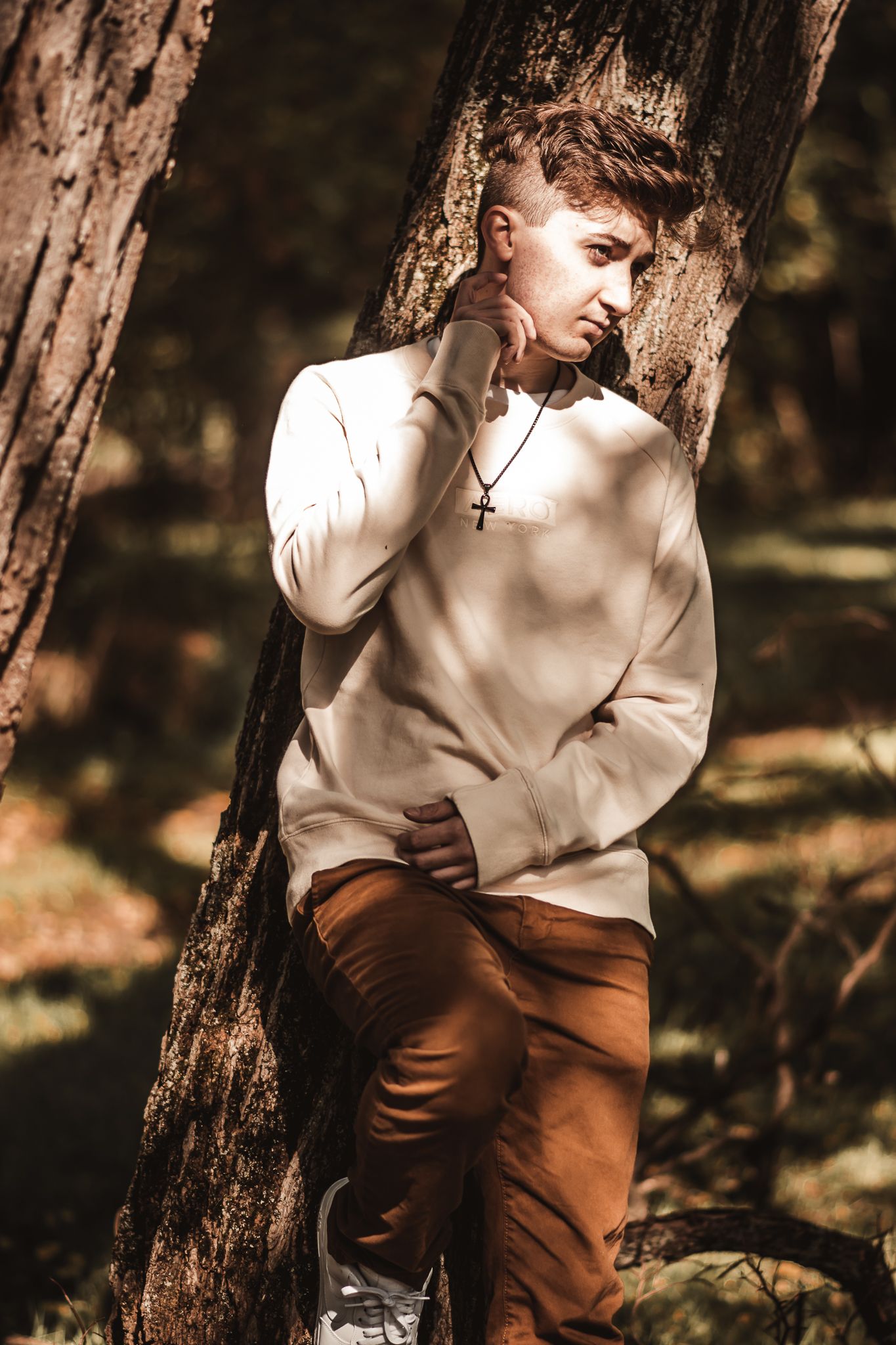 Portrait of a young person in beige sweater leaning against a tree trunk in natural lighting.