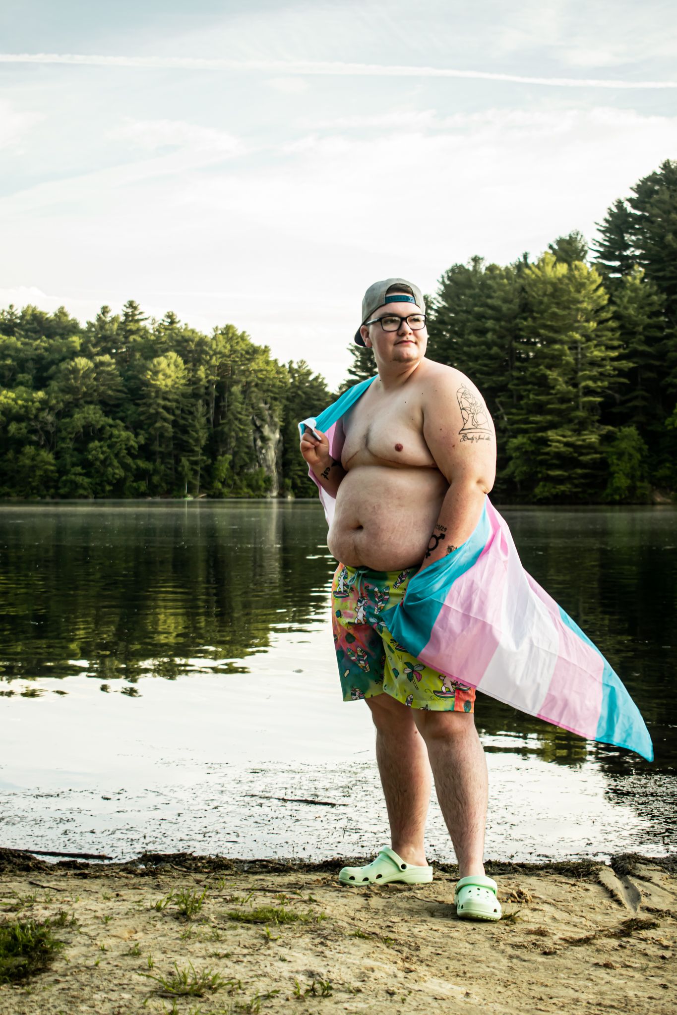 A person at a lakeside beach holding a trans pride flag and posing confidently.