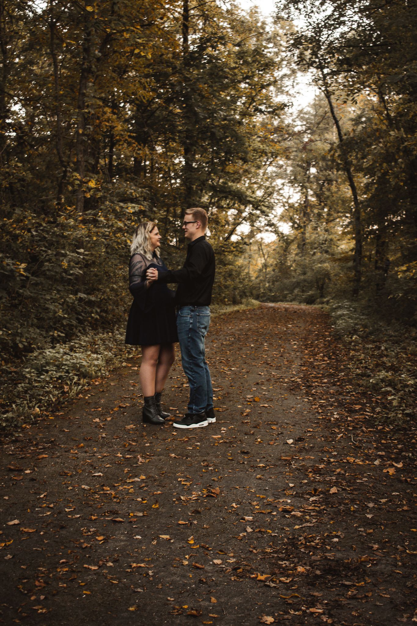 A romantic couple standing on a leaf-covered forest path during autumn.