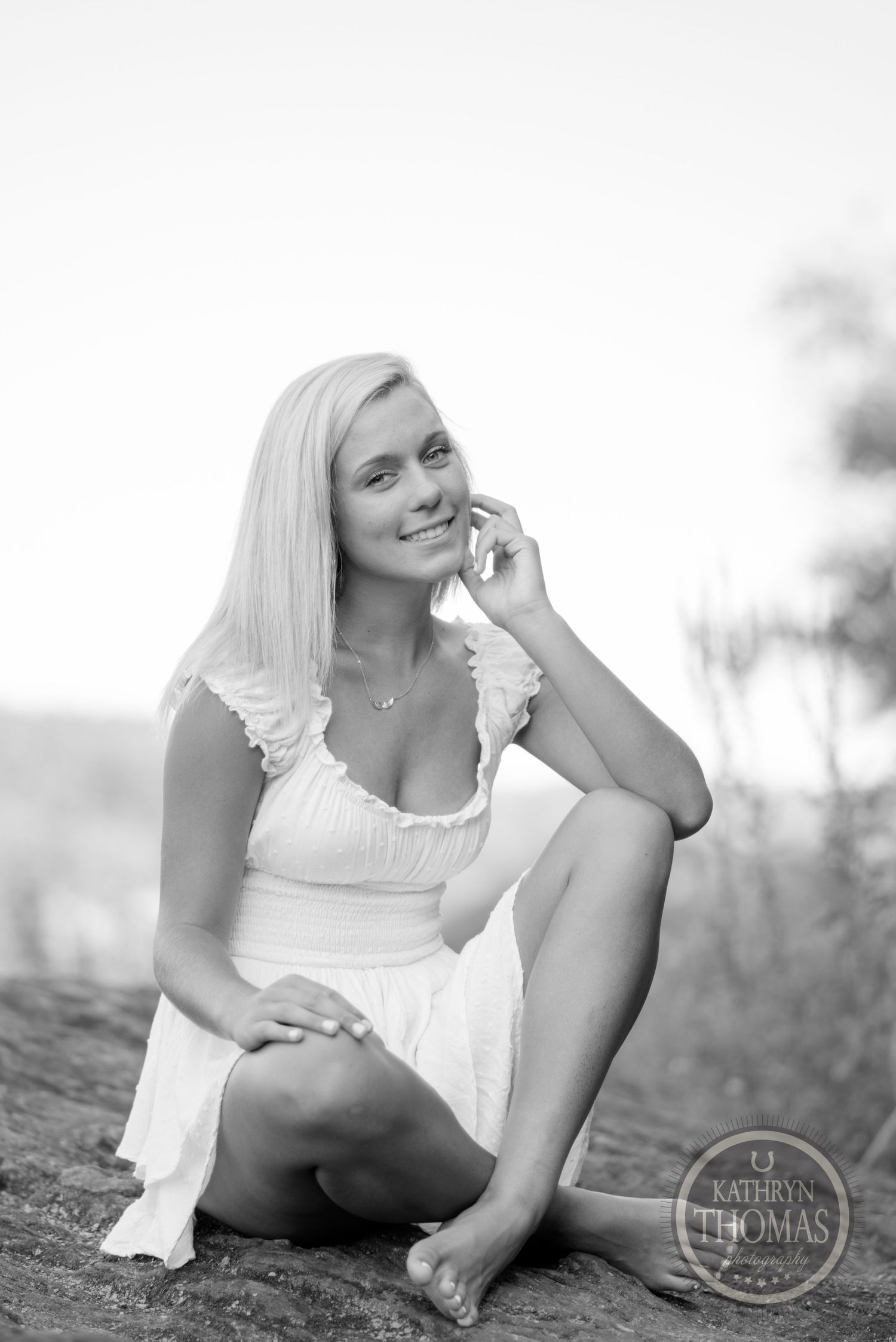 Black and white portrait of someone in a white dress sitting casually on rocky ground.