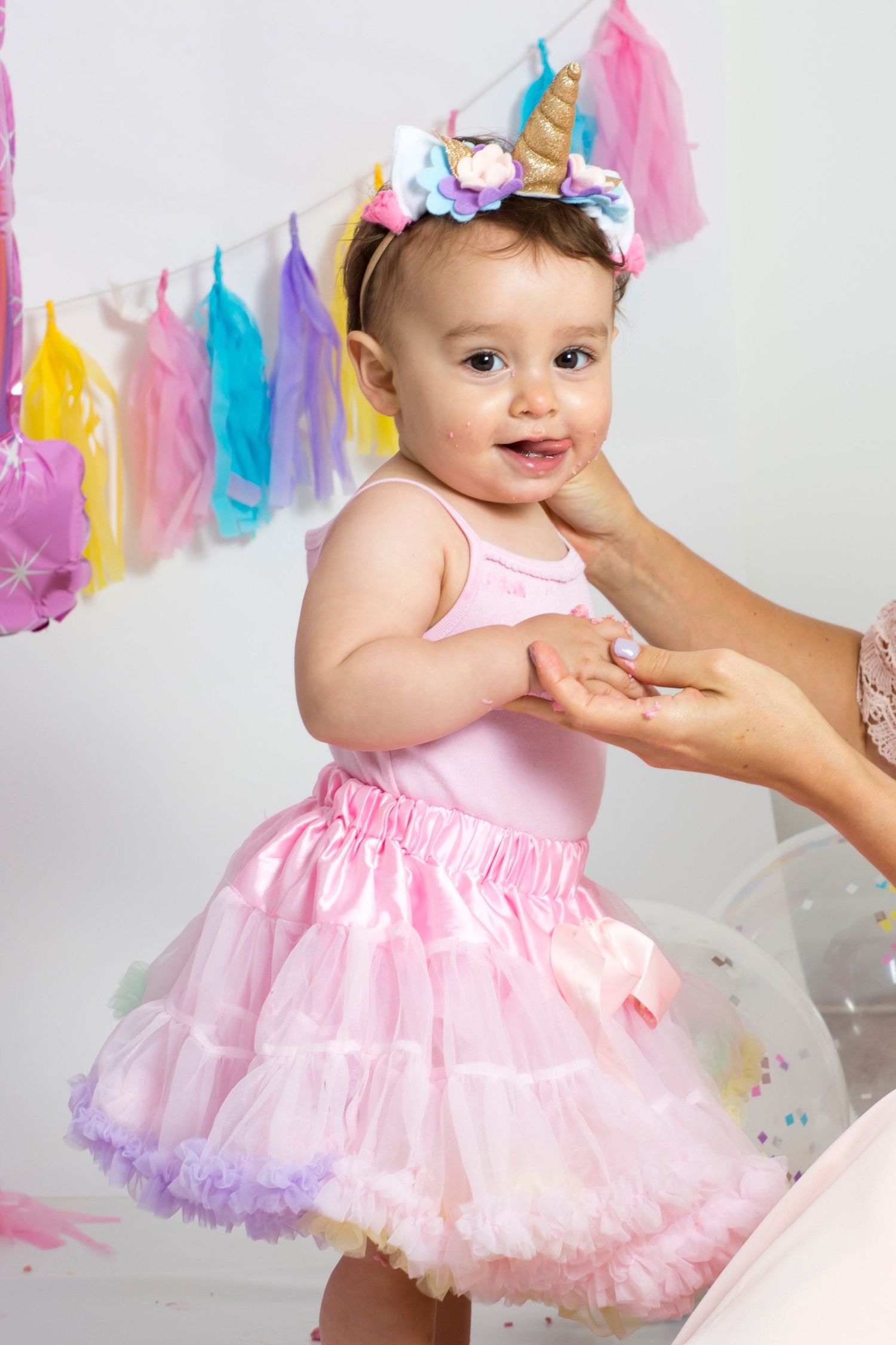 A baby wearing a unicorn headband and pink tutu dress at a colorful birthday celebration.