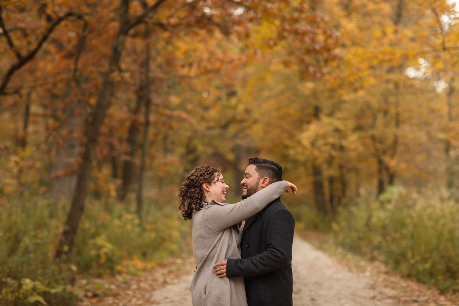 A romantic autumn engagement photo of a couple embracing on a tree-lined path with golden fall foliage.