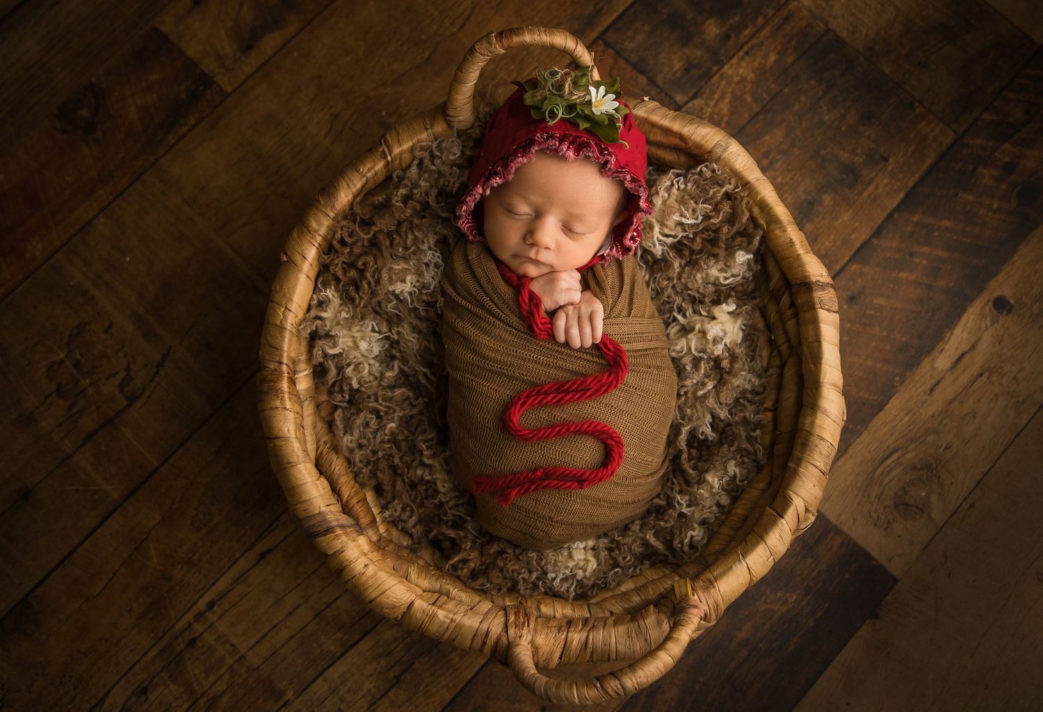Sleeping newborn wrapped in red outfit lies in rustic wooden bowl with holiday decorations.