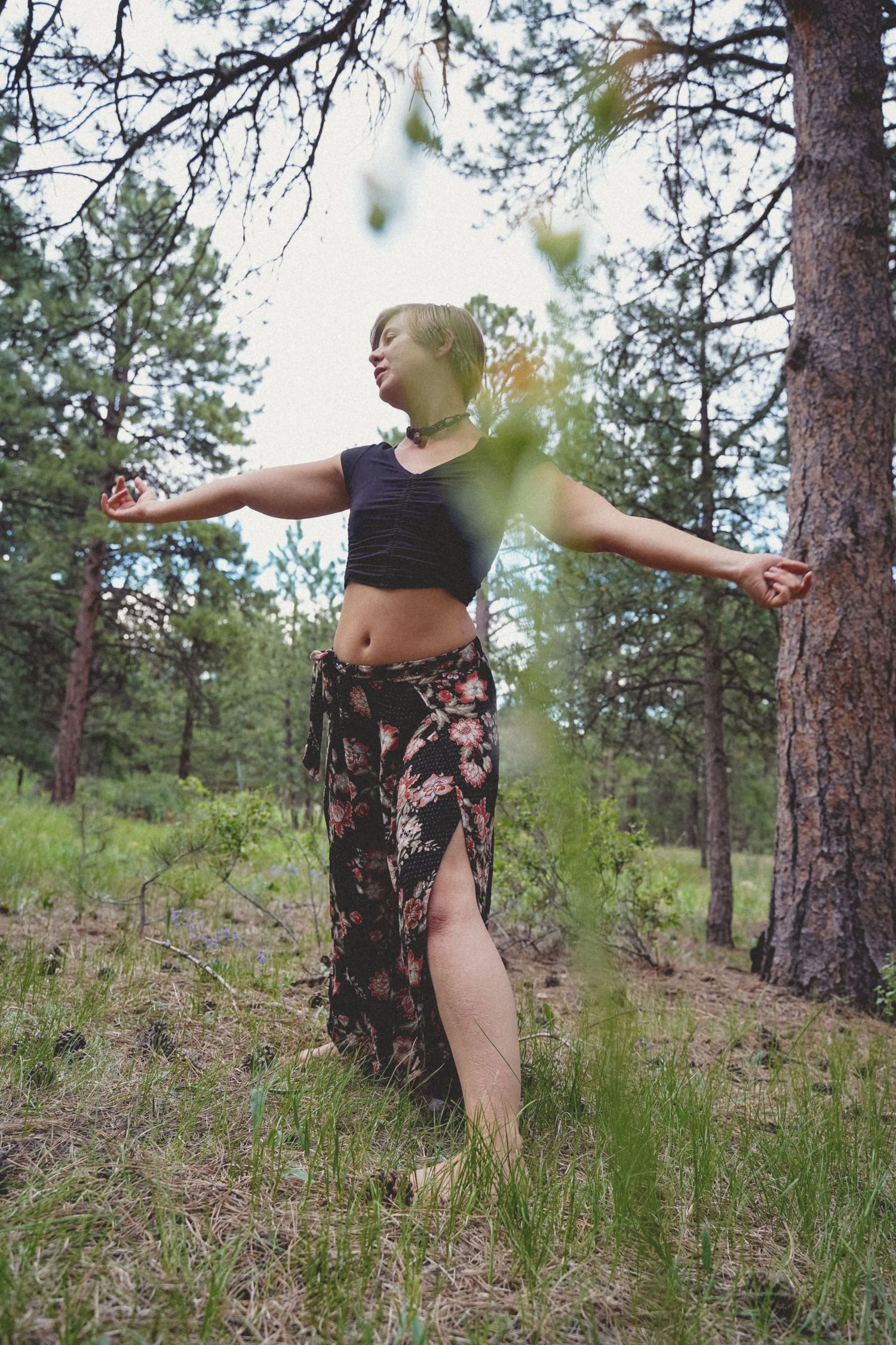 Dancing figure in crop top and patterned skirt among pine trees in forest setting.