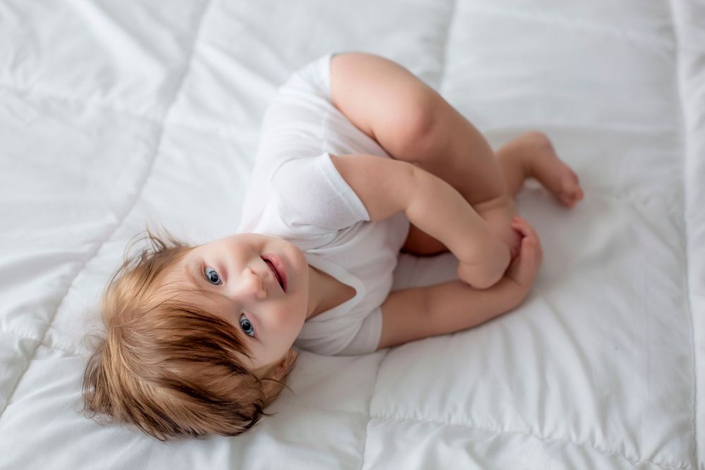 6 month old baby boy laying on white blanket with white onesie looking up at camera.