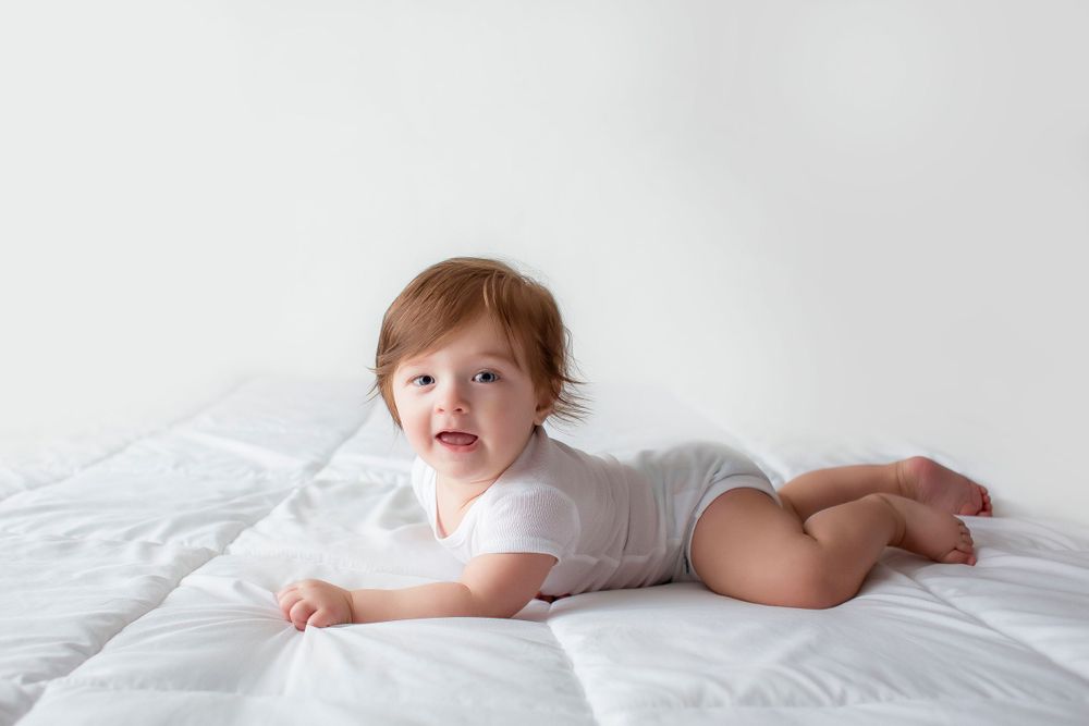 6 month old baby boy laying on white blanket in a white onesie doing tummy time.
