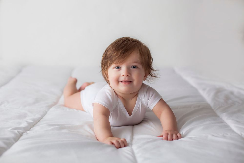 6 month old baby boy in a white onesie doing tummy time on white blanket.