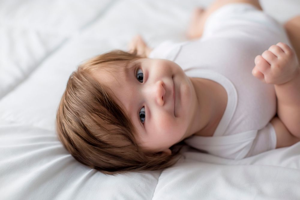 6 month old baby boy on white blanket in white onesie laying on back looking at camera smiling.