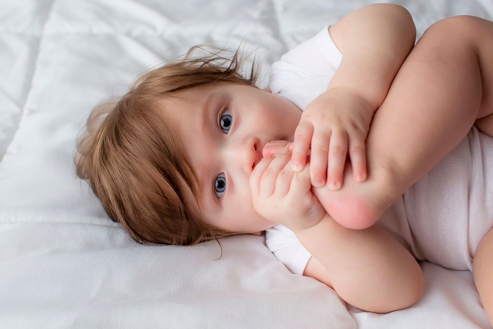 6 month old baby boy on a white blanket eating his toes.