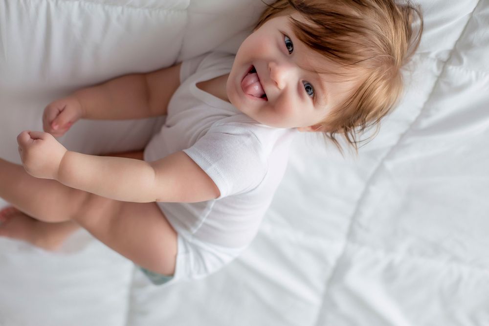 6 month old baby boy laying on back with white blanket looking at camera sticking out tongue.